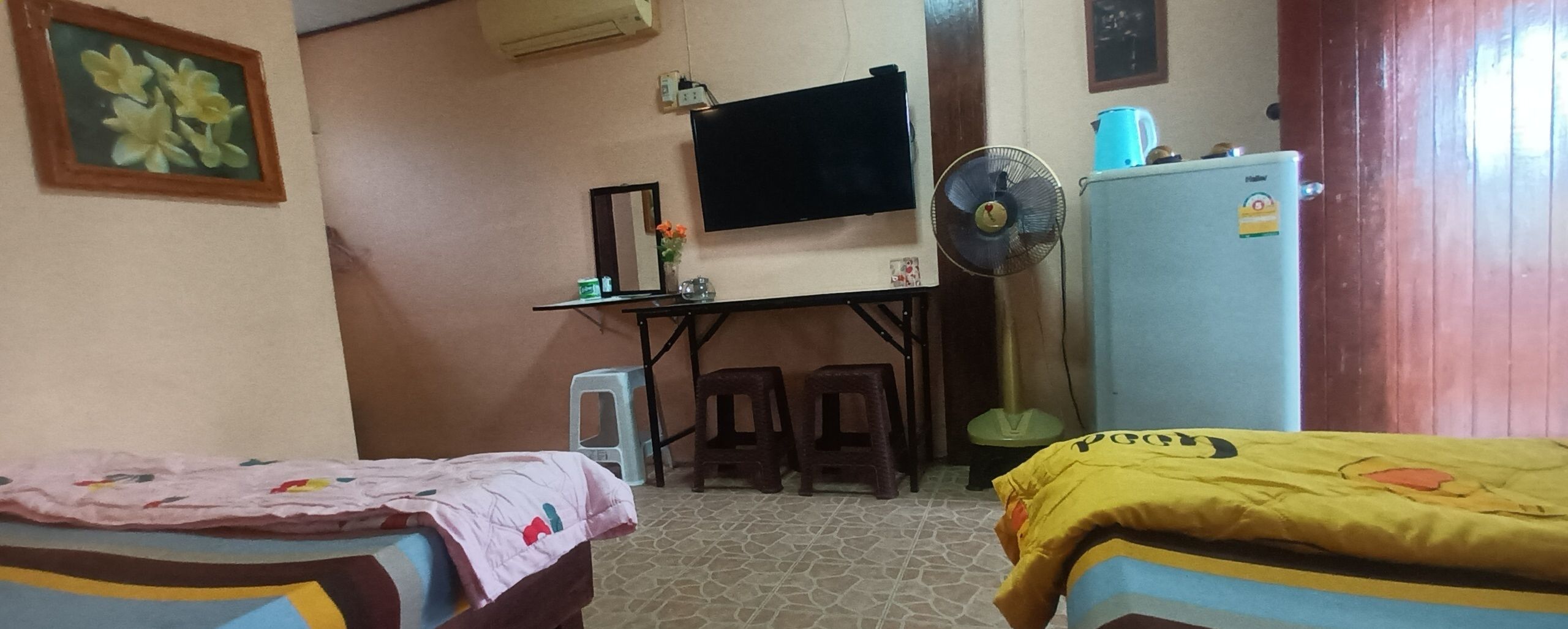 A bedroom with two beds, a desk with a TV, a fan, and a refrigerator.