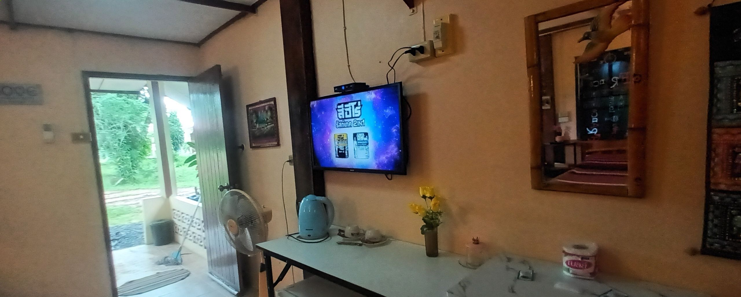 Huoneessa on televisio, jossa näkyy ohjelma, tuuletin, vedenkeitin ja pieni pöytä, jonka päällä on maljakko täynnä kukkia.