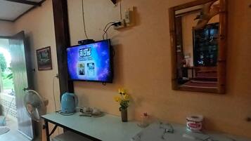 A room with a television displaying a program, a fan, a kettle, and a small table with a vase of flowers.