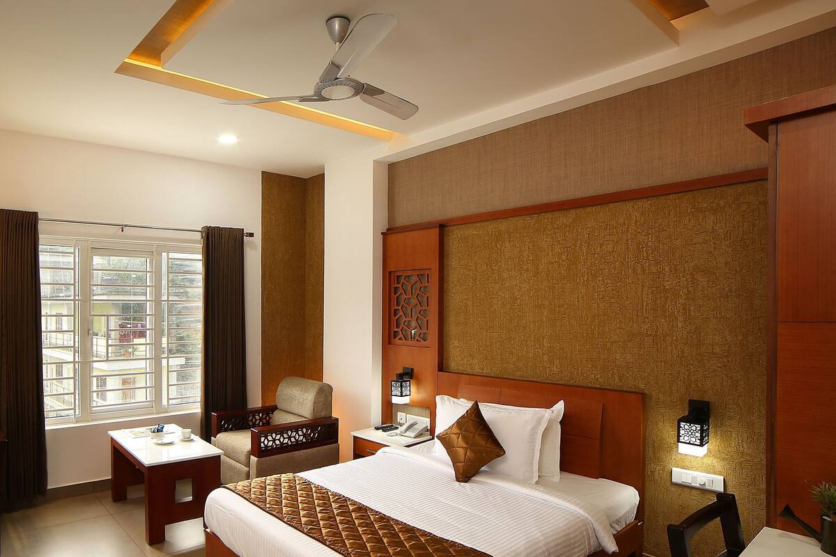 A hotel room with a bed, a desk, a chair, a TV, a window with curtains, a ceiling fan, and a wall-mounted light fixture.