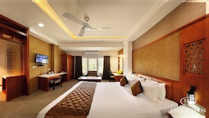 A hotel room with a bed, desk, chair, television, and a ceiling fan. - Kgees- HillTown Hotel (Idukki)