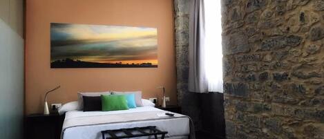 A hotel room with a bed, a nightstand, a painting of a sunset, a stone wall, and a window with curtains.