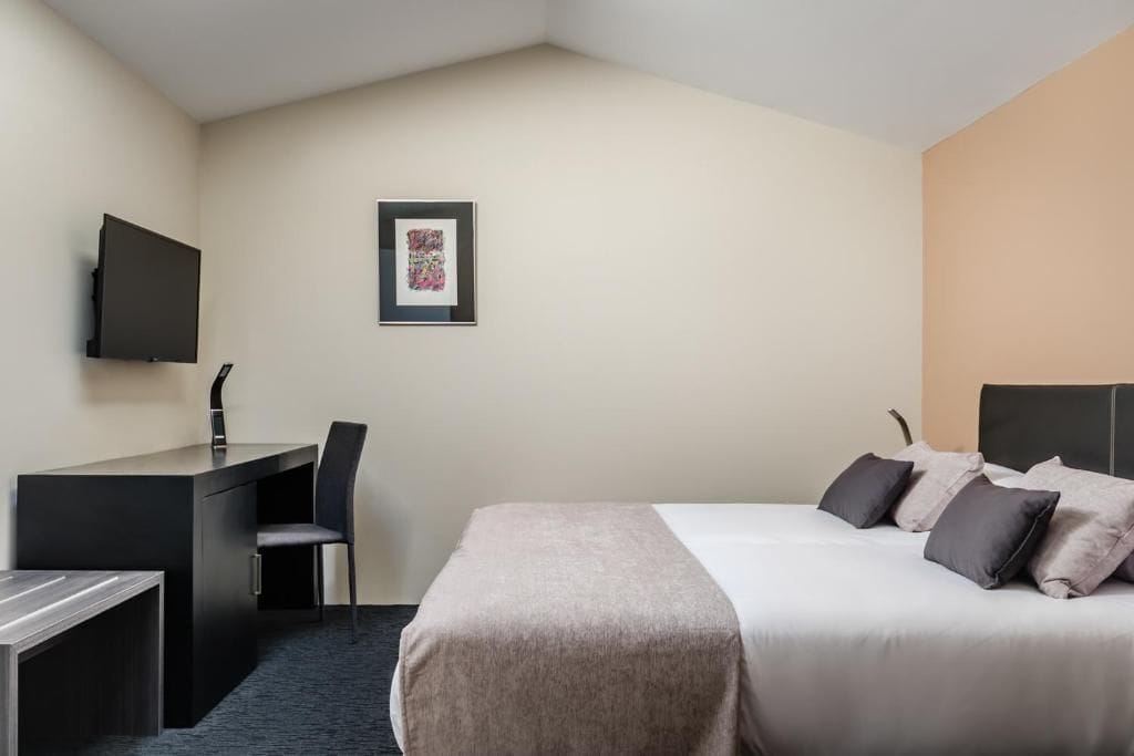 Ein Hotelzimmer mit einem Bett, einem Schreibtisch, einem Sessel, einem Fernseher und einem gerahmten Bild an der Wand.