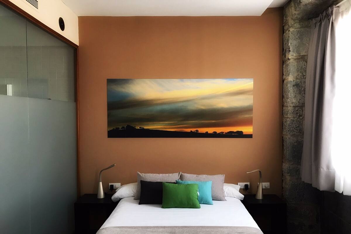 Une chambre d’hôtel avec un grand lit, un tableau représentant un coucher de soleil, un mur en pierre et une fenêtre avec des rideaux.