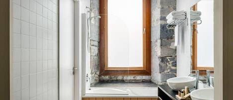 Un baño con pared de piedra, ventana con marco de madera, bañera y dos lavamanos.