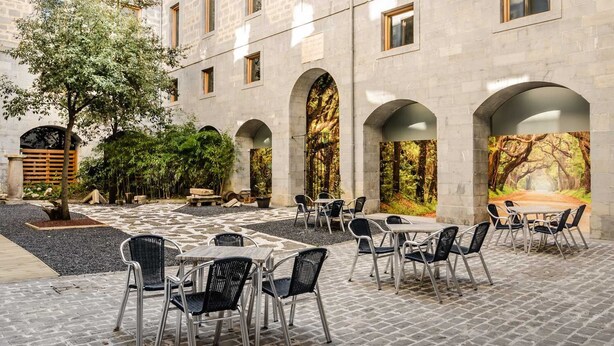 Un patio al aire libre con suelo de piedra, varias mesas y sillas, y vistas a un bosque a través de ventanas arqueadas.