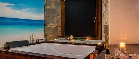 A spa room with a large bathtub, a stone wall, a view of the ocean, and a table with candles and flowers.