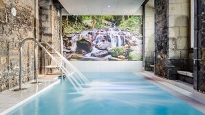 Una piscina interior con un mural de cascada, paredes de piedra y una escalera de metal.