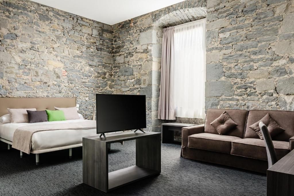 Een kamer met een stenen muur, een bed, een bank, een televisie op een standaard, en een raam met gordijnen.
