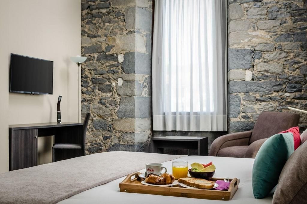 Ein Hotelzimmer mit Steinwand, einem Bett, einem Schreibtisch mit Fernseher und einem Frühstückswagen mit Toast, Obst und Saft.