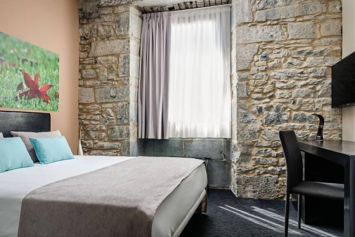 Une chambre avec un mur en pierre, un lit recouvert d’une couverture beige, un bureau avec une chaise et une fenêtre avec des rideaux.