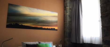 Une chambre d’hôtel avec un grand lit, un tableau représentant un coucher de soleil, un mur en pierre et une fenêtre avec des rideaux.