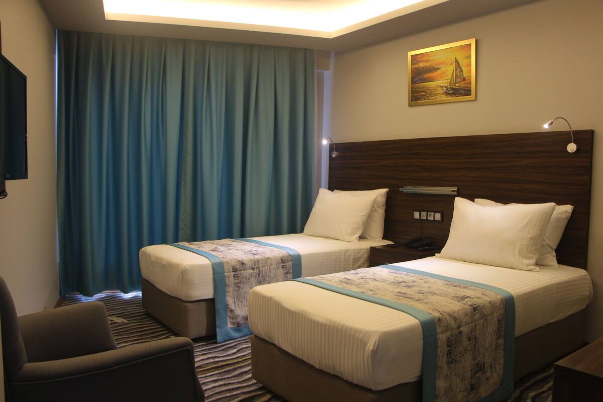 A hotel room with two beds, a desk, a chair, a painting, and a curtain.