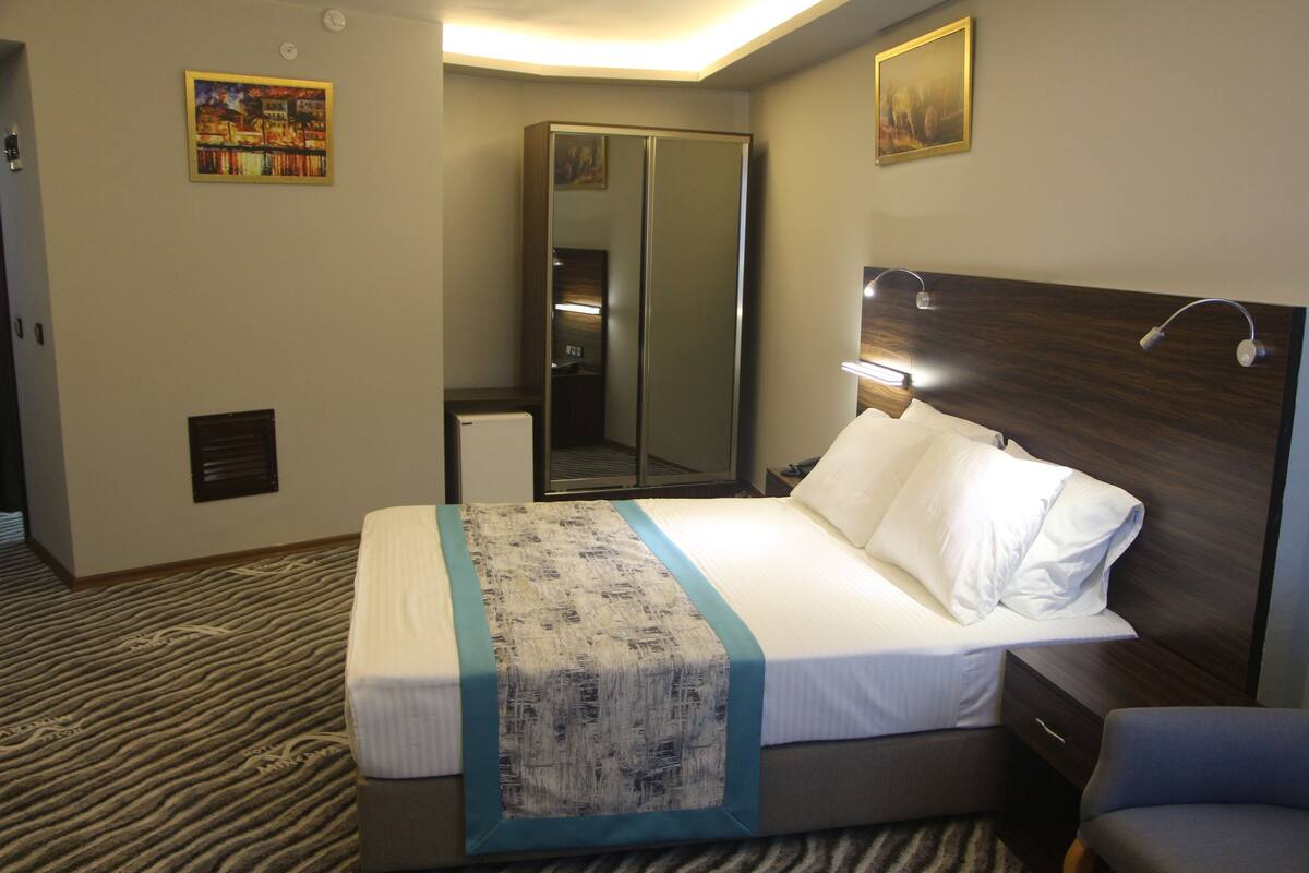A hotel room with a bed, bedside table, nightstand, lamp, mirror, wardrobe, and a carpeted floor.
