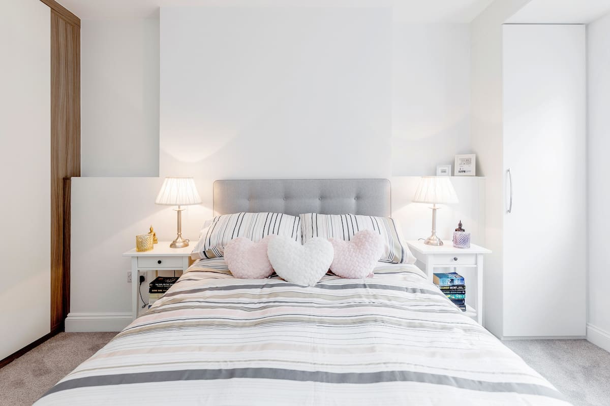 Un dormitorio ordenado con una colcha a rayas, dos lámparas de noche y almohadones con forma de corazón.