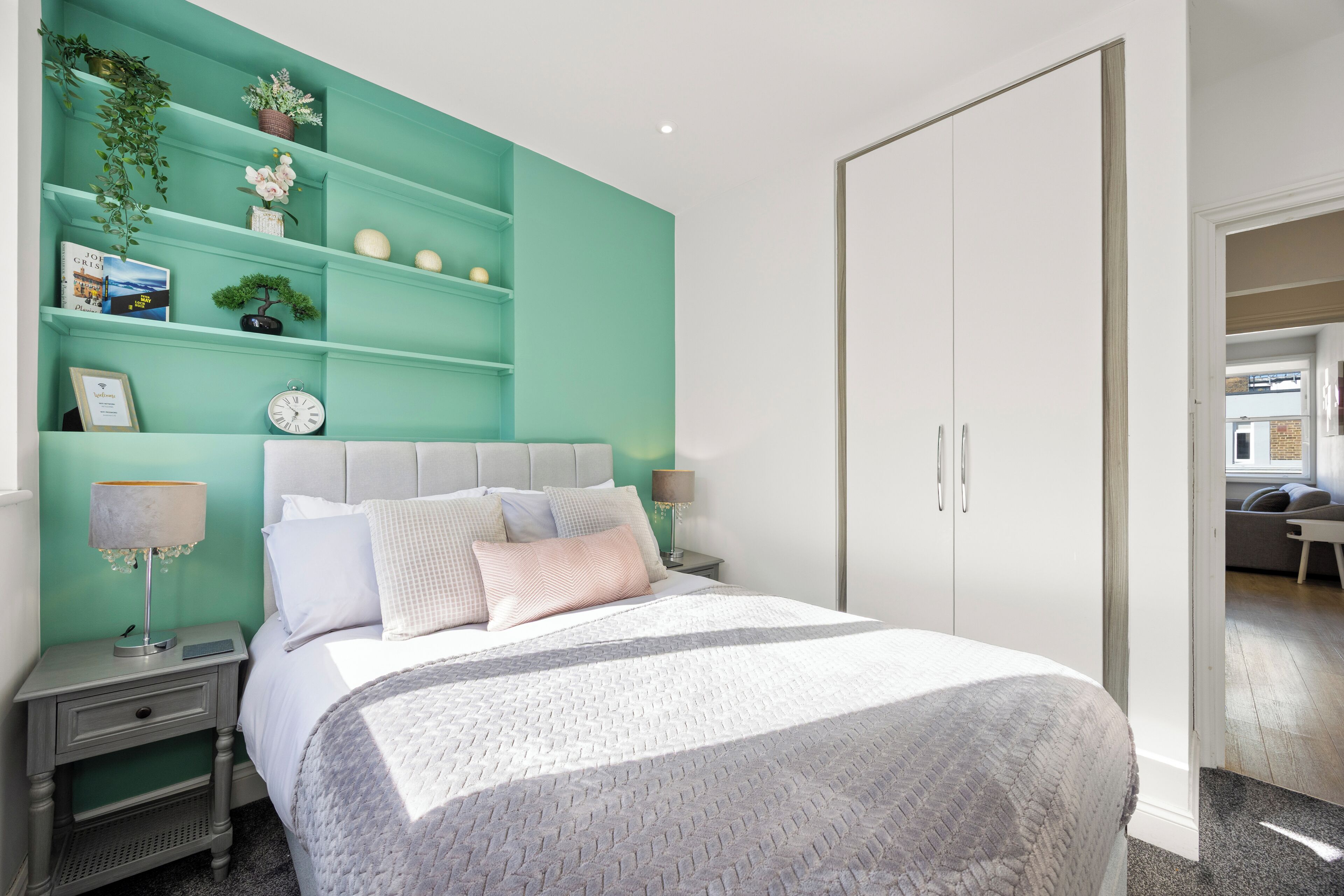 A modern bedroom with a teal accent wall, a large bed with a patterned bedspread, a nightstand with a lamp, and a bookshelf with decorative items.