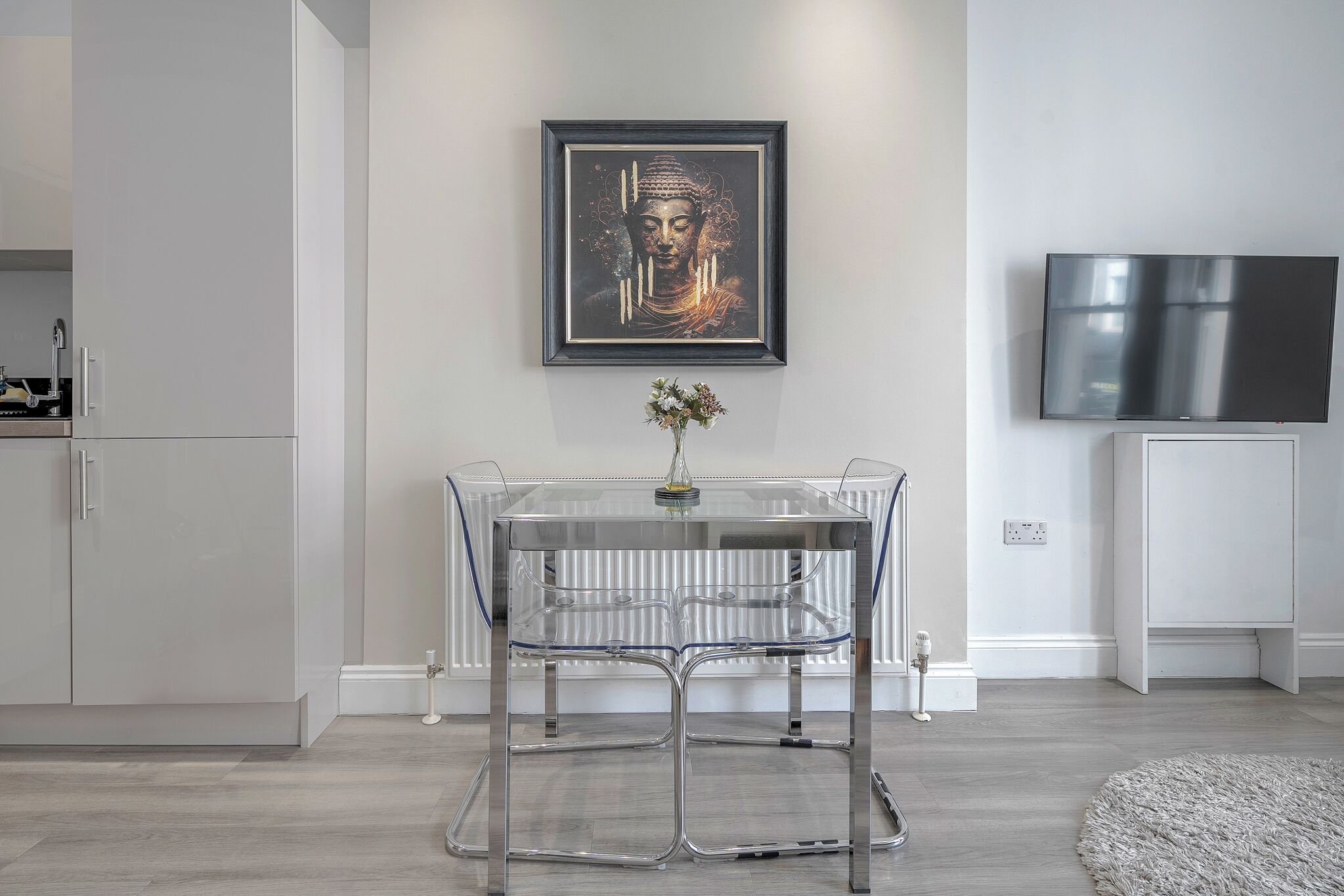 A modern kitchen with a glass-top table, a framed artwork above it, and a flat-screen TV on the wall.