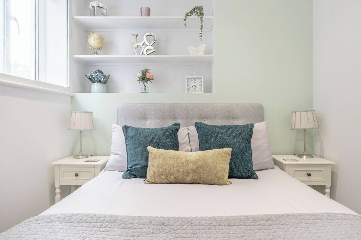 Un dormitorio ordenado con cama, dos mesitas de noche, un estante con adornos y una ventana.