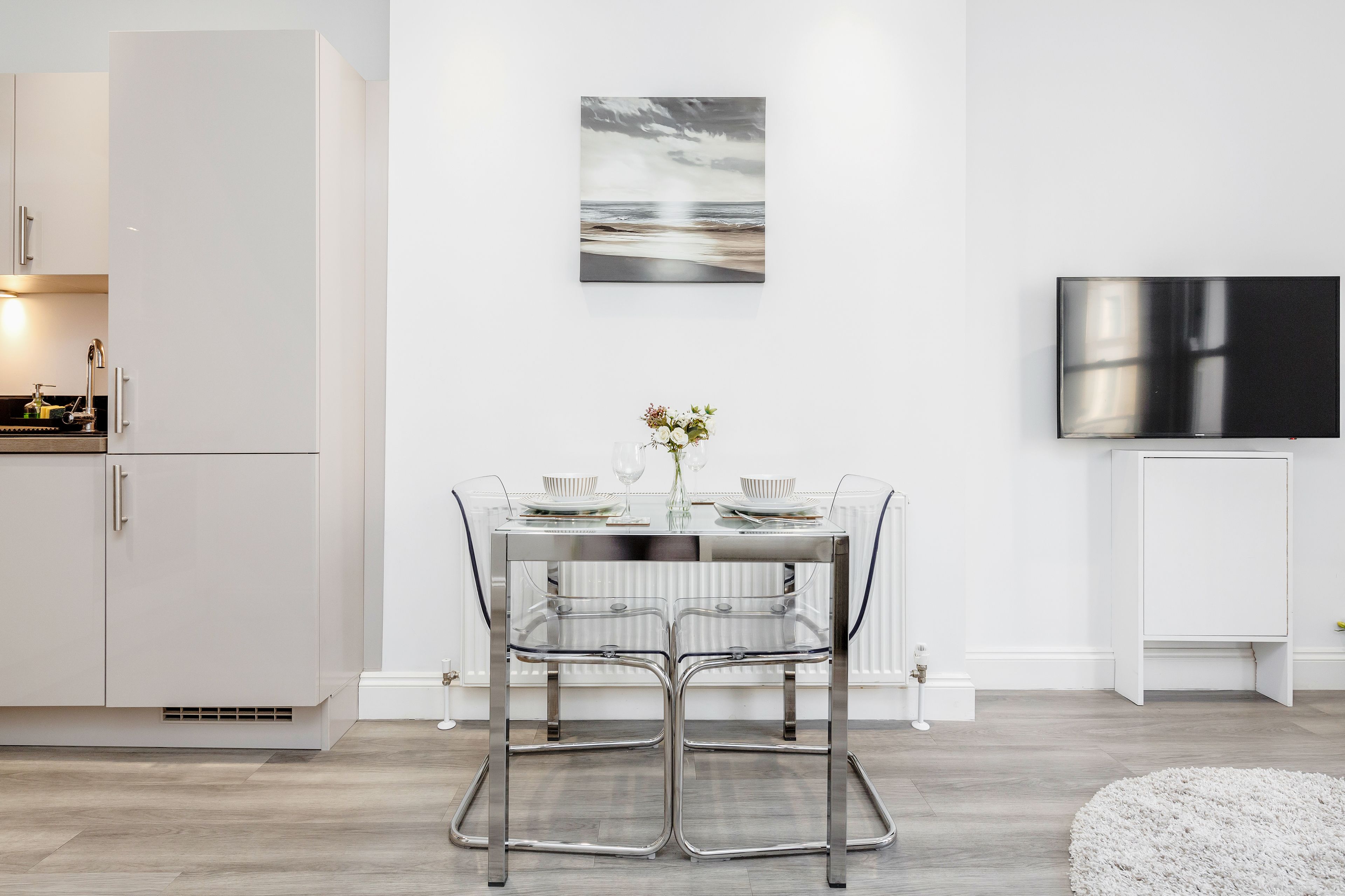 A modern kitchen with a dining area featuring a glass table and chairs, a flat-screen TV, and a framed artwork on the wall.