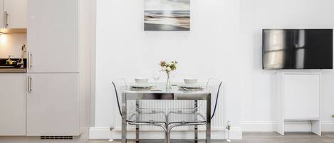 A modern kitchen with a dining area featuring a glass table and chairs, a flat-screen TV, and a framed artwork on the wall.