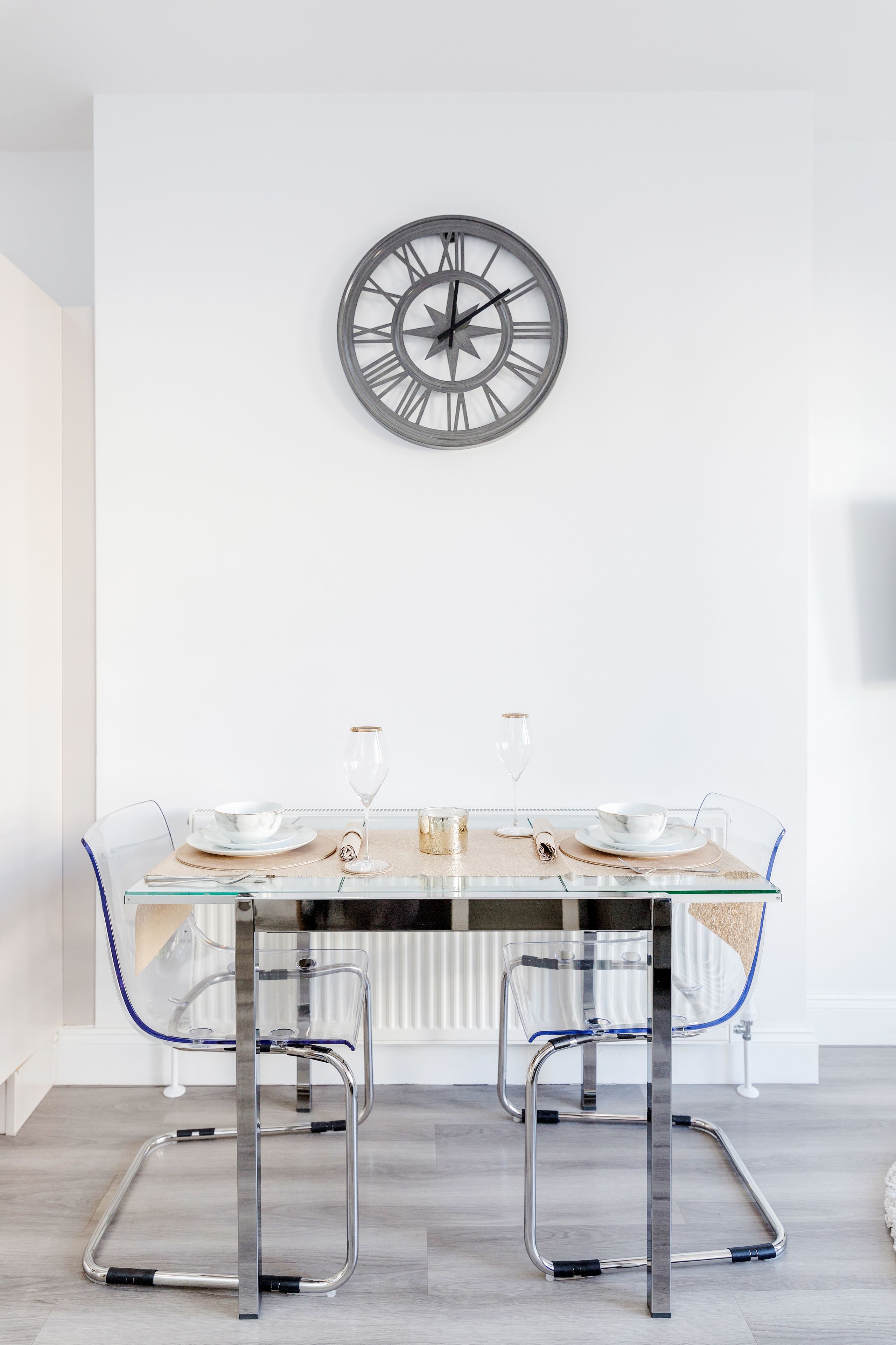 A dining area with a glass table, two chairs, a clock, and a radiator.