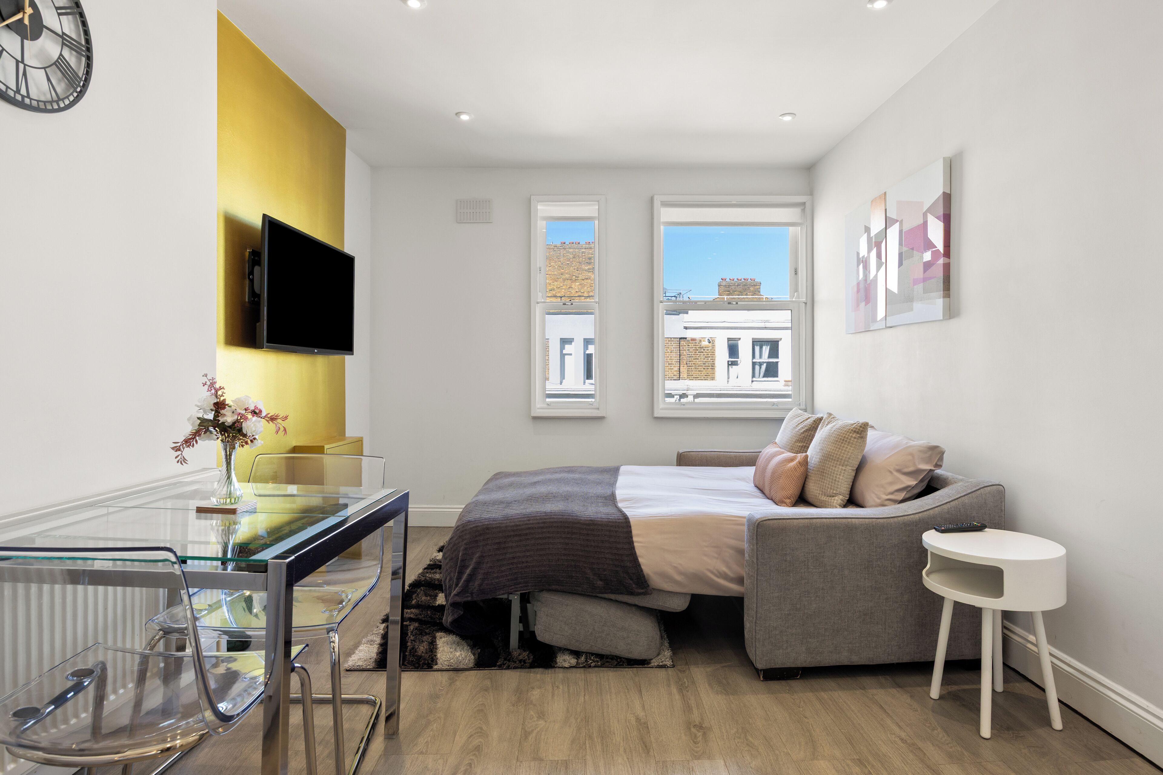 A modern bedroom with a bed, a glass desk, a TV, and a view of buildings through windows.