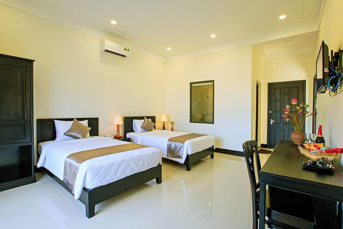 A hotel room with two beds, a desk, a chair, a dining table, a vase with flowers, and a painting on the wall.