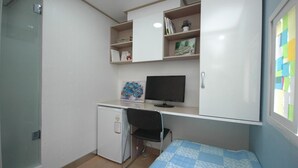 A compact bedroom with a desk, a small refrigerator, a bed with a blue quilt, and a window with a view of colorful notes. - LotteTower Rainbow House (Seoul)