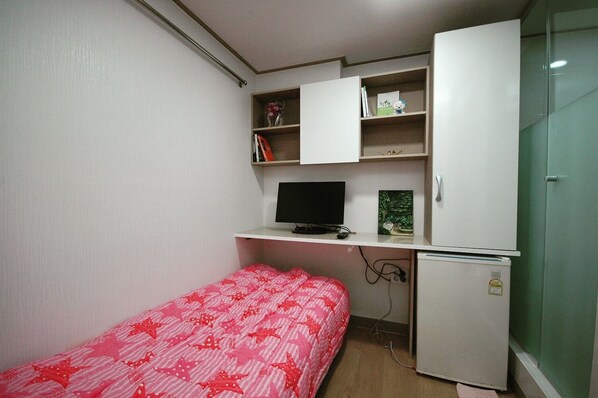 A small, compact room with a bed, a desk, a computer, and a refrigerator.
