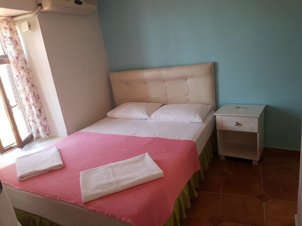A neatly made bed with a pink bedspread and white pillows, a white bedside table with a drawer, and a window with floral curtains. - Assos Dr No Pansiyon (Ayvacik)