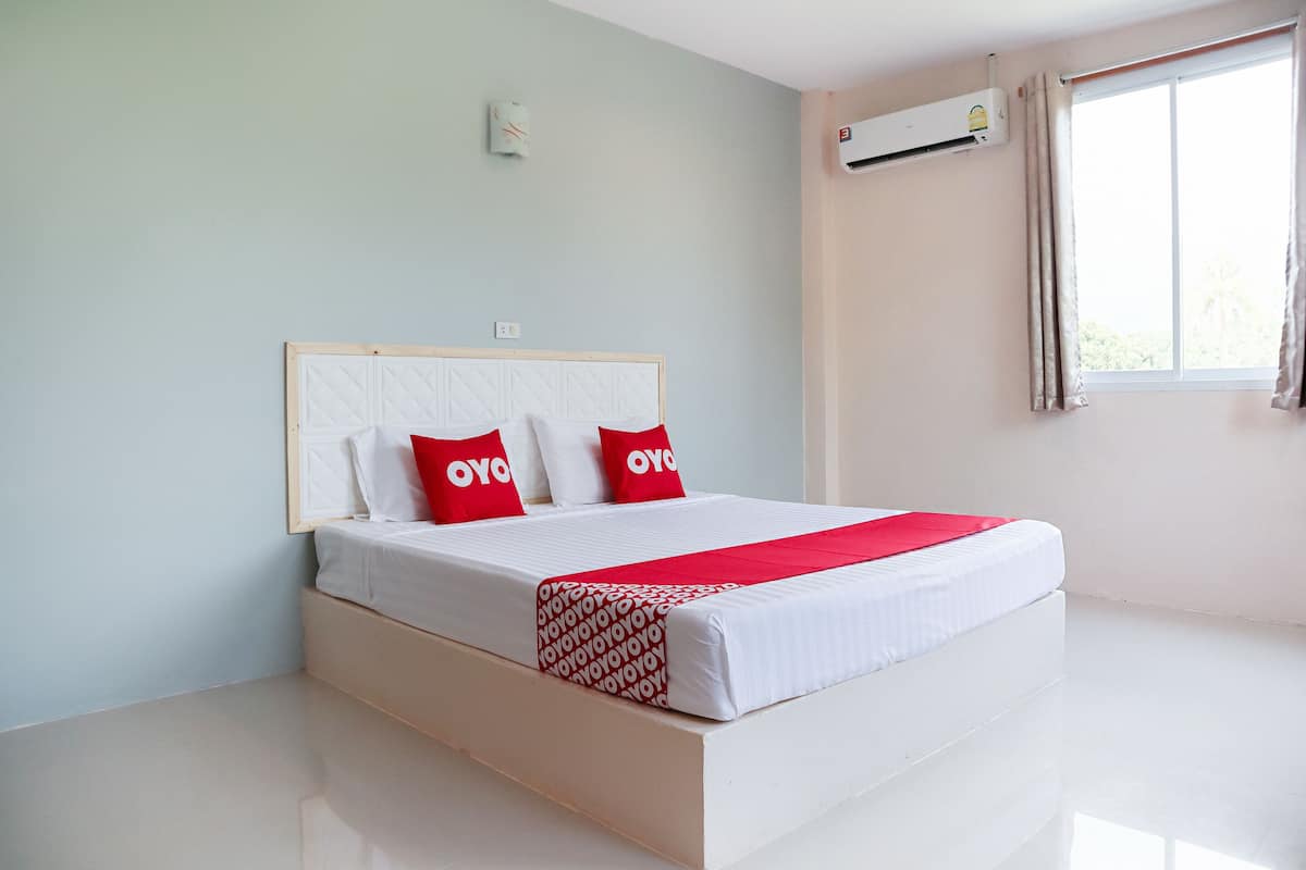 Un lit bien fait, avec du linge de lit blanc et des oreillers rouges, une fenêtre avec des rideaux et un appareil de climatisation fixé au mur.