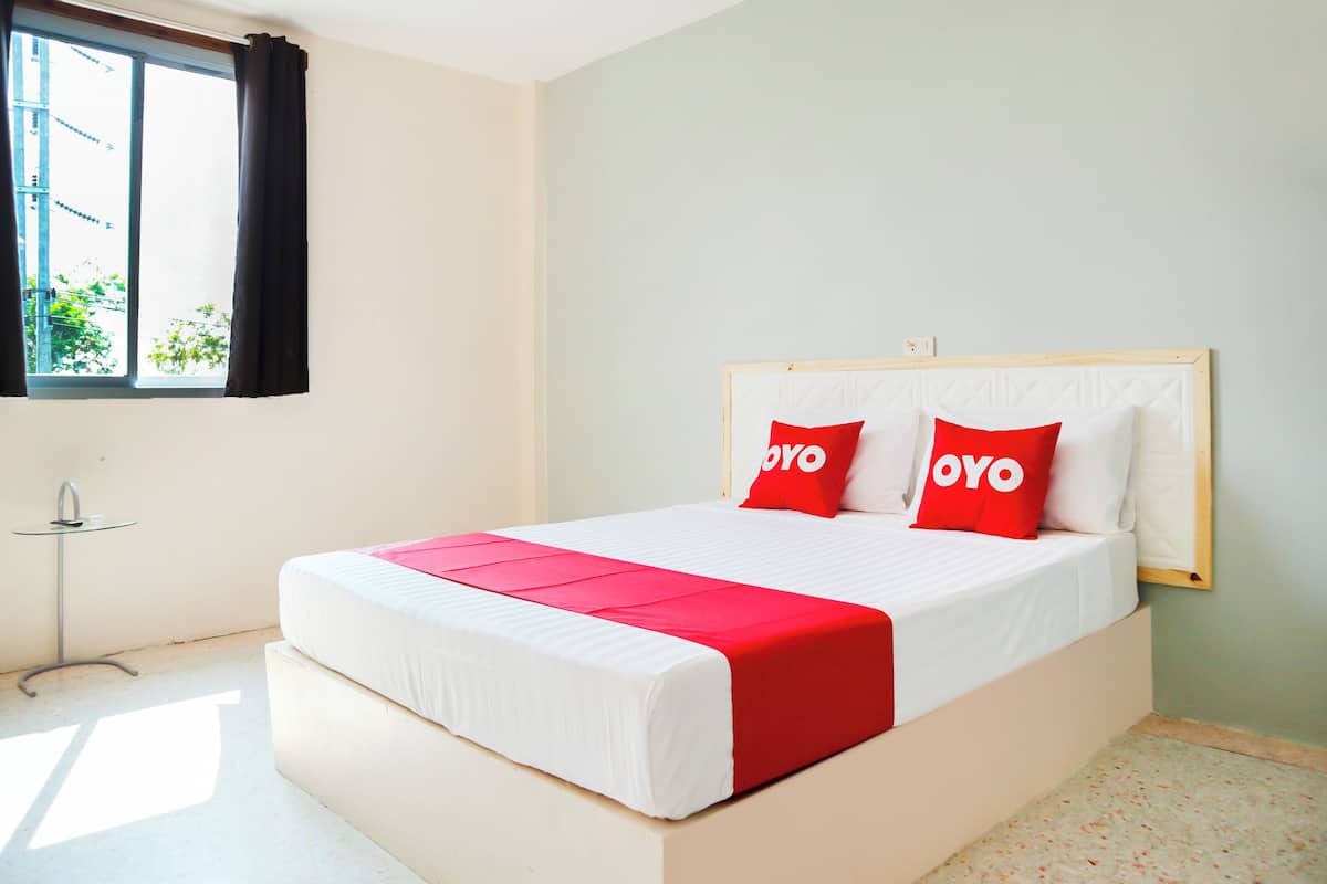 Un lit bien fait, recouvert d’une couette blanche et rouge, avec deux oreillers rouges sur lesquels est inscrit le mot « OYO », et une fenêtre avec des rideaux noirs.