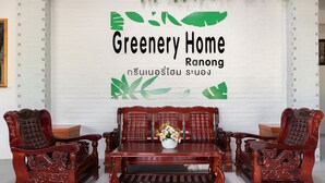 A traditional wooden sofa set with intricate carvings, a small table with a vase of flowers, and a white brick wall with green leaf patterns and text. - OYO 674 Greenery Home (Ranong)