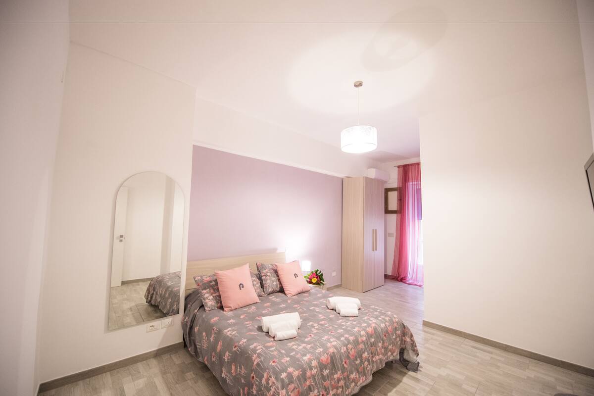 A bedroom with a bed, a mirror, a nightstand, and a door leading to another room.