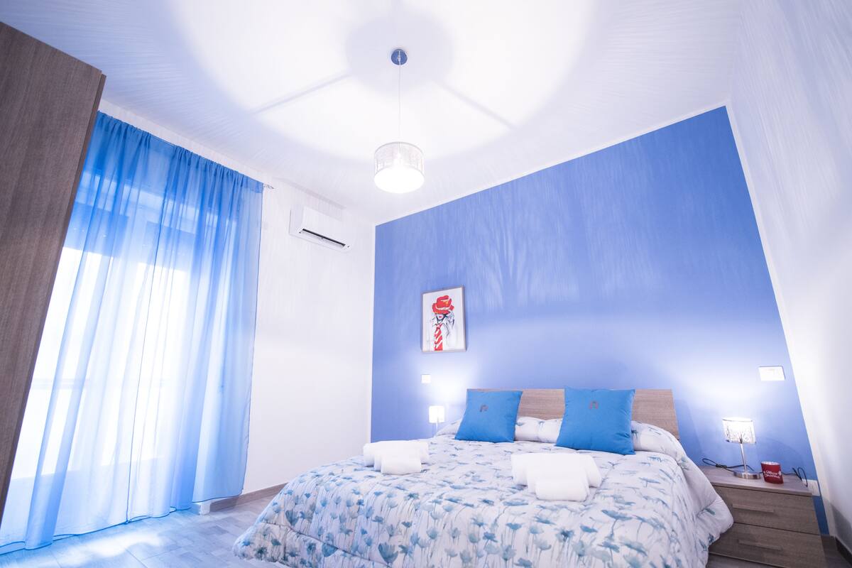 A bedroom with a large bed, blue walls, and a window with sheer curtains.