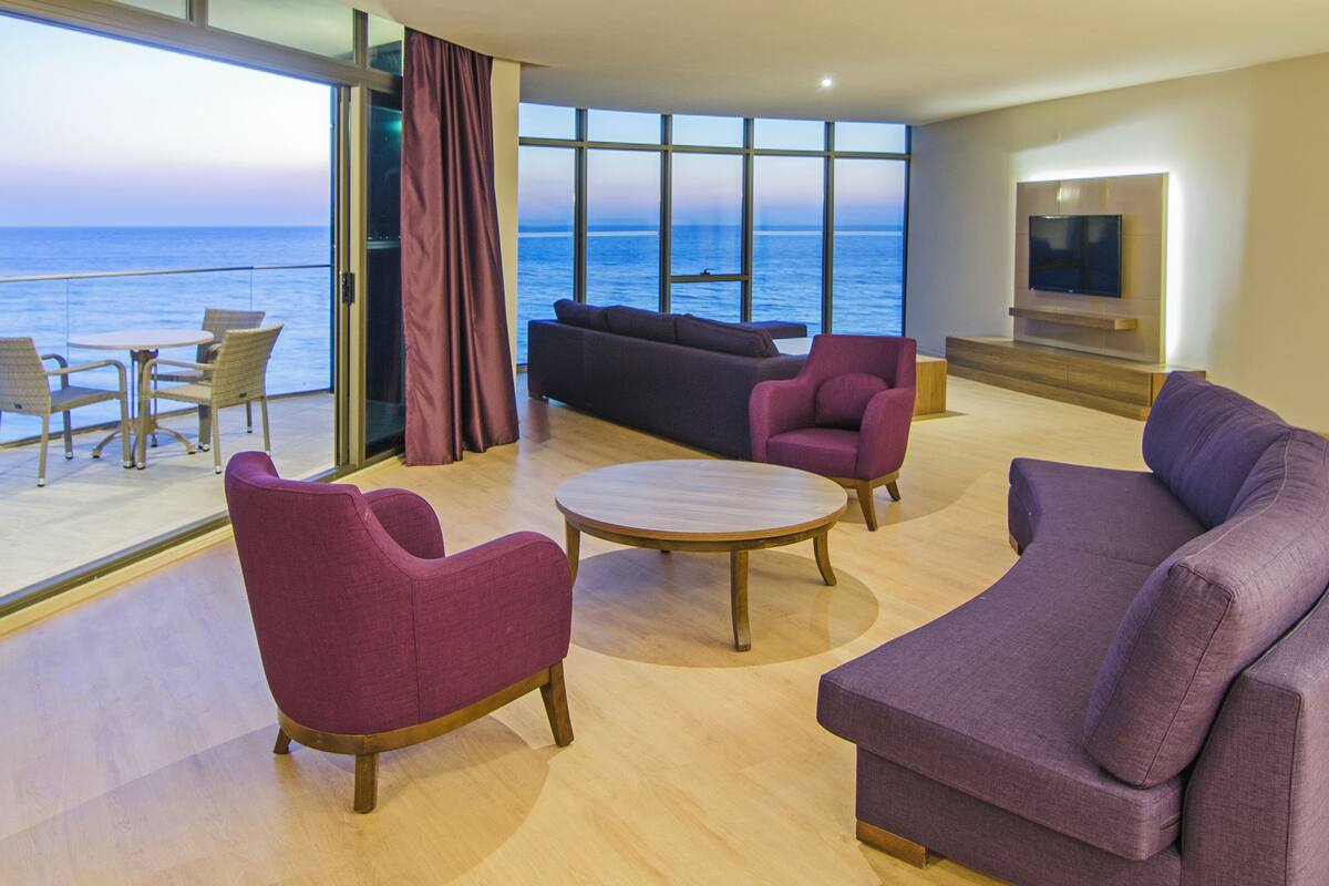 Una sala de estar moderna con un gran ventanal con vistas al mar, amueblada con sofás morados, sillones y una mesa de centro.