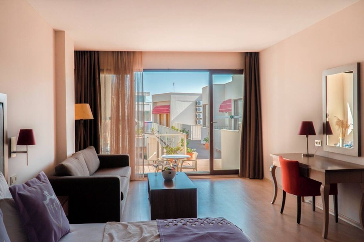 Habitación de hotel con sofá, escritorio, silla, mesa de centro y ventana grande con vistas a edificios.