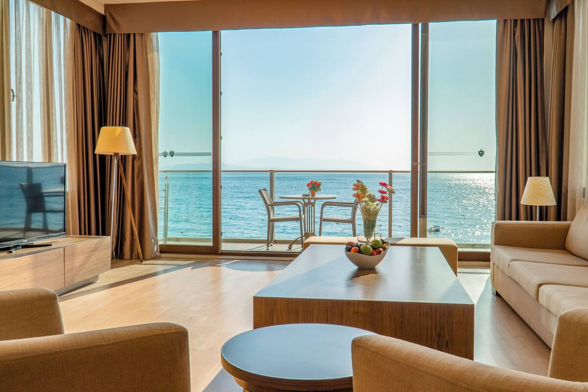 Habitación de hotel moderna con un balcón amplio, televisión de pantalla plana, sofá, mesita de centro y mesa de comedor con sillas.