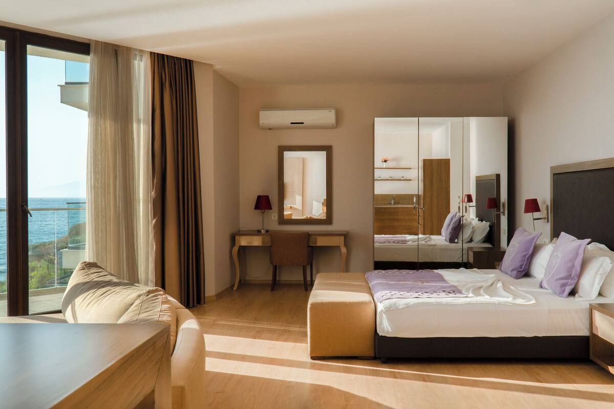 Habitación de hotel con cama, escritorio, silla, mesita de noche, espejo y vista al mar.