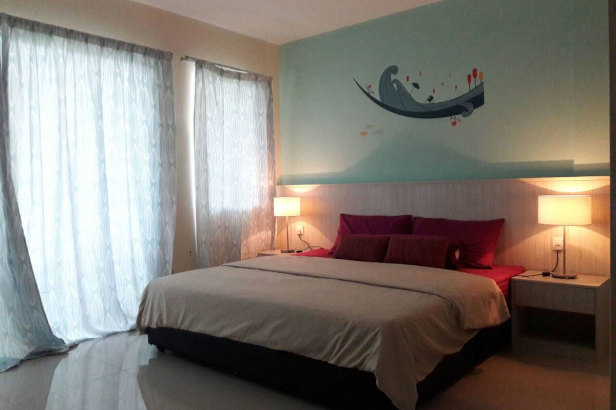 Ein Schlafzimmer mit einem Bett, zwei Nachttischen mit Lampen, einer Wand mit einem Wandbild und Vorhängen an den Fenstern.