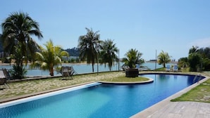 A swimming pool with a wooden deck and lounge chairs, surrounded by palm trees and a view of the sea. - Marina Island Apartment (Lumut)