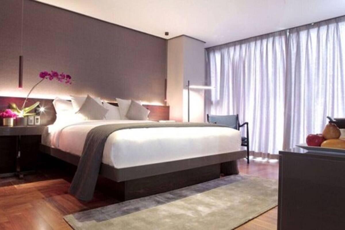 Una habitación de hotel moderna con una cama grande, una mesita de noche con lámpara, una silla y vistas al exterior a través de la ventana.
