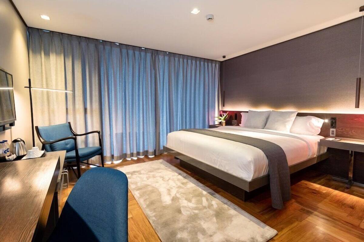 Una habitación de hotel moderna con una cama grande, un escritorio con silla, televisión y un ventanal con cortinas.