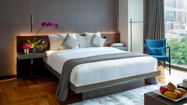 A modern hotel room with a large bed, a nightstand with a vase of flowers, a blue chair, and a view of the city. - Ramada Encore Makati (Makati City)