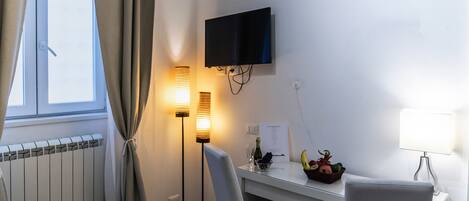 A hotel room with a desk, chair, television, floor lamp, and a window with curtains.