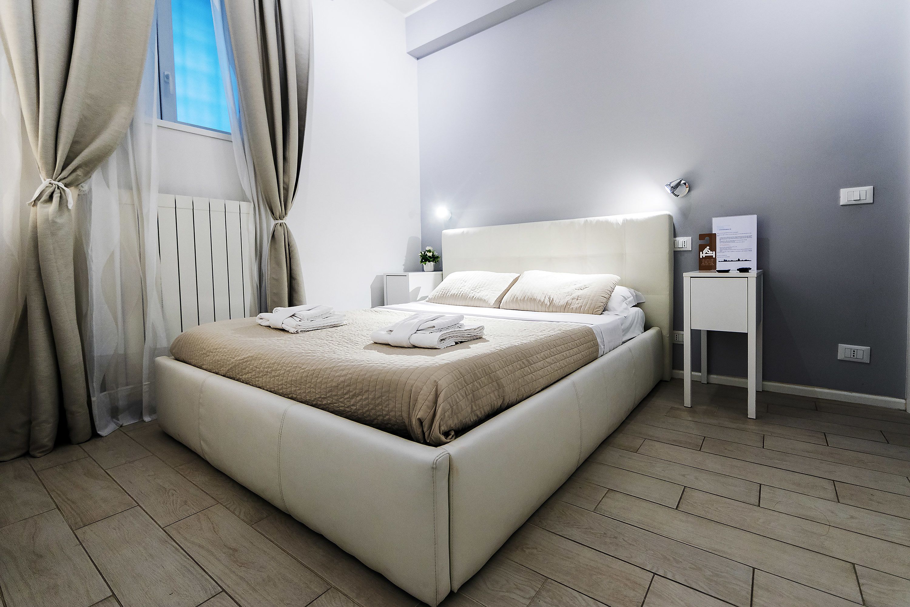 Una camera da letto moderna con un letto grande, tende bianche, un comodino con una lampada e una piccola pianta.