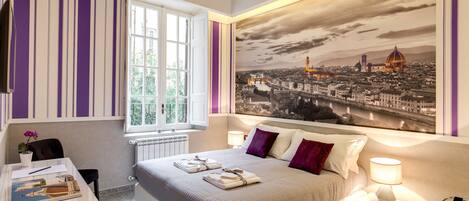 A modern hotel room with a large bed, a desk with a computer, a window with curtains, and a cityscape mural on the wall.