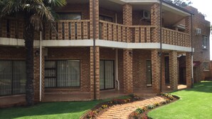 A two-story brick building with a balcony, large windows, and a paved garden area with flowers.