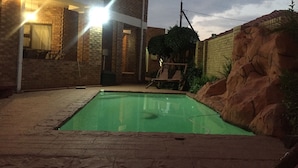 A brick house with a swimming pool, a wooden swing, and a large rock formation.