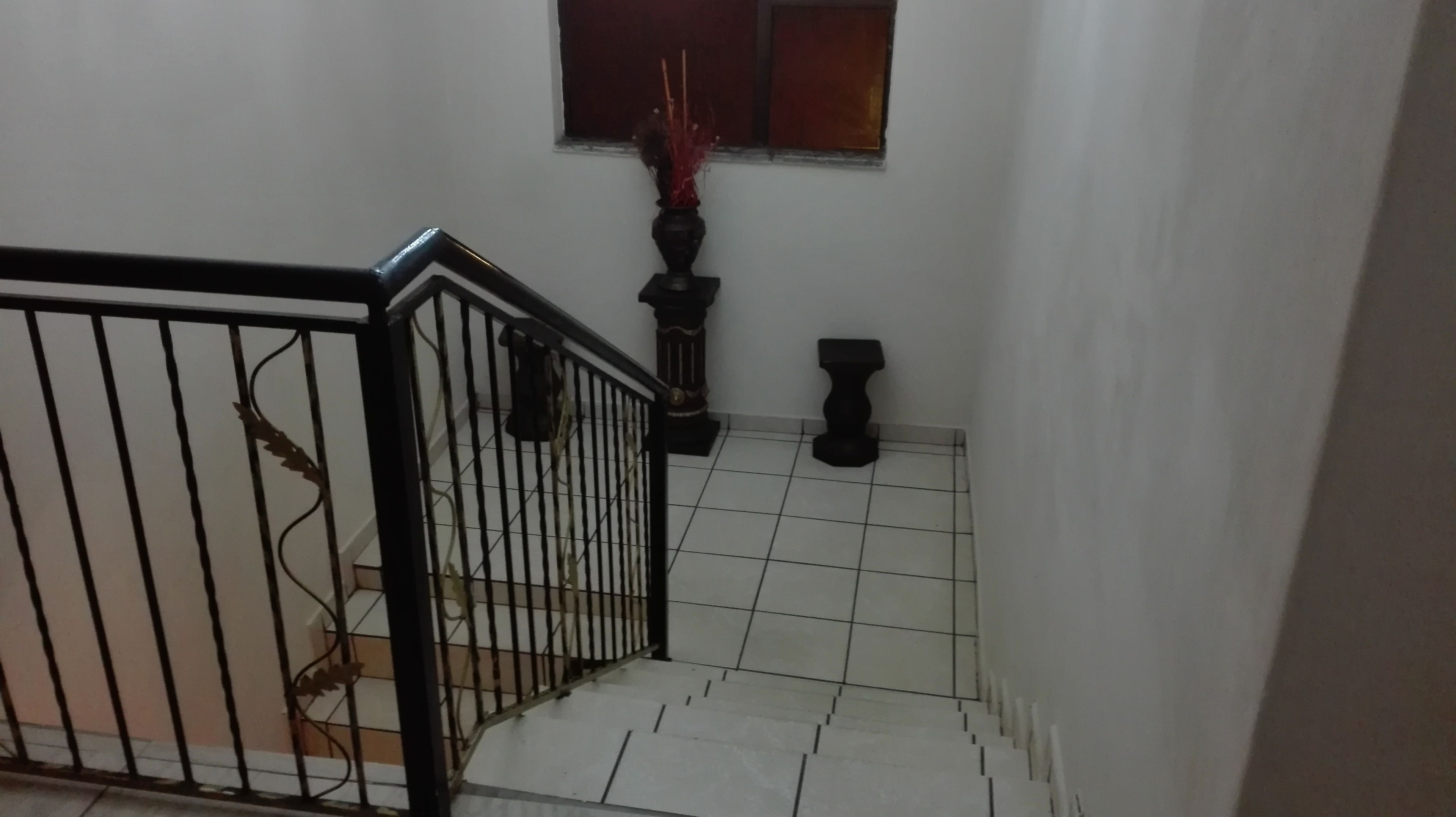 Una escalera con barandilla de metal negro, suelo de baldosas blancas y un pequeño ventanal con un adorno rojo.
